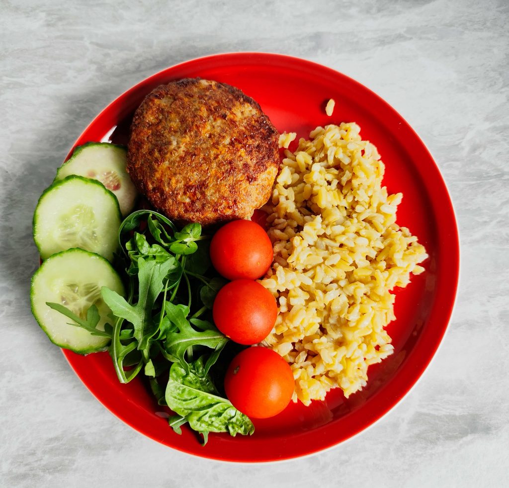 Balanced meal with chicken patty, brown rice, cucumber slices, cherry tomatoes and fresh greens on a red plate
